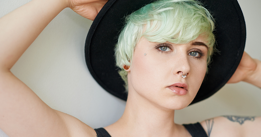 A white person with more feminine features, greenish hair, wearing a black tank top and a black hat, has their arms raised above their head and gazes at the viewer.